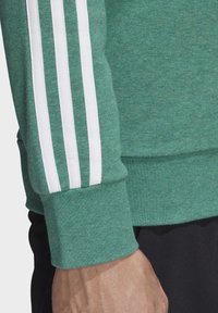 Manche de sweat-shirt vert avec trois bandes blanches et un poignet, porté par une personne avec une main visible et un pantalon noir.
