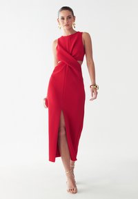 Red maxi dress with a fitted bodice, twist detail at the waist, and side slit. Made of textured fabric, featuring a sleek, sleeveless design.