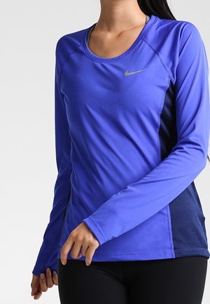 Femme portant un t-shirt de sport Nike bleu à manches longues avec des panneaux latéraux en mesh noir et un legging noir, posant avec les bras détendus.