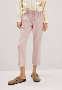 Pantalones cortos de color rosa claro con cintura elástica y cordón, que cuentan con un único bolsillo lateral y dobladillos sin terminar, combinados con zuecos marrones.