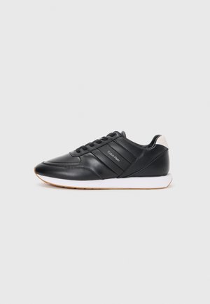 Sneakers sintetici neri con una texture liscia, caratterizzati da strisce orizzontali, una suola bianca e un accento beige sul tallone.