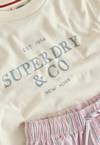 T-shirt crème avec logo Superdry & Co, les inscriptions "EST 1954" et "New York", associé à un short à rayures rose et blanc avec taille élastique.