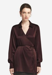 Burgundy satinomslagen blus med långa puffärmar, en sjalkrage och en knuten midja, med en jämn yta och en v-ringning.