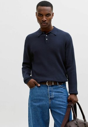 Mężczyzna w ciemnoniebieskim prążkowanym polo z długim rękawem i niebieskich dżinsach, trzymający brązową skórzaną torbę prawą ręką.