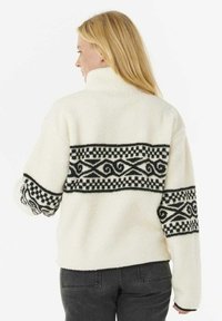 Crème-kleurige fleece jack met een hoge kraag, voorzien van een zwart patroonontwerp op de rug en mouwen. Structuur en gezellig.