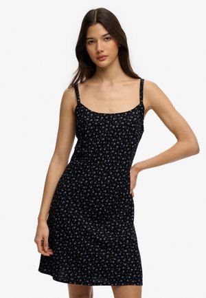 Jeune femme aux longs cheveux noirs portant une robe noire sans manches à petites fleurs violettes, posant avec une main sur la hanche.