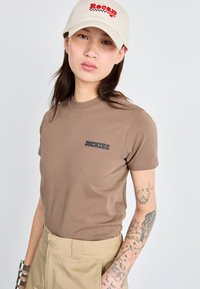 T-shirt in cotone marrone con logo "Dickies" nero, abbinato a pantaloni khaki e un cappellino beige con testo ricamato rosso.
