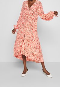 Šaty s červenobílým vzorem, dlouhými rukávy, výstřihem do V a asymetrickým lemem. Hladká látka s volánkovými detaily na manžetách rukávů.