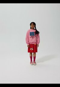 Pige med flettet hår iført lyserød sweatshirt med blå gitterdesign, rød nederdel med ko grafik, lyserøde sokker og røde sneakers, stående på hvid baggrund.