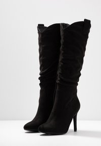 Botas de ante negras hasta la rodilla con punta afilada, tacón alto de aguja y diseño arrugado. Cuentan con un cierre lateral y un ligero pliegue en la parte superior.
