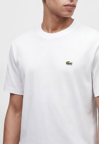 Junge Person trägt ein schlichtes weißes T-Shirt mit kurzen Ärmeln und kleinem grünem Krokodil-Logo auf der linken Brust.