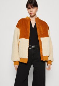 Chaqueta de pana marrón claro y forro de crema con puños y cuello de canalé, con cierre de cremallera y bolsillos laterales. Llevada sobre una blusa negra.