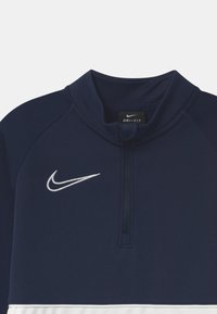 Sudadera de Nike Dri-FIT azul marino con logotipo swoosh blanco en el lado izquierdo del pecho y collar corto con cremallera por encima de una franja horizontal blanca.