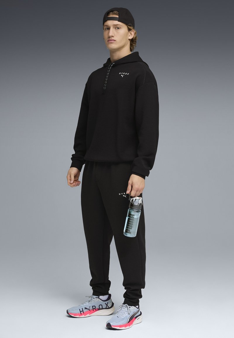 Νεαρό άτομο που φοράει μαύρο φόρμα HYROX και καπέλο, κρατώντας ένα διαφανές αθλητικό μπουκάλι νερού, στέκεται μπροστά σε φόντο με γκρι διαβάθμιση.