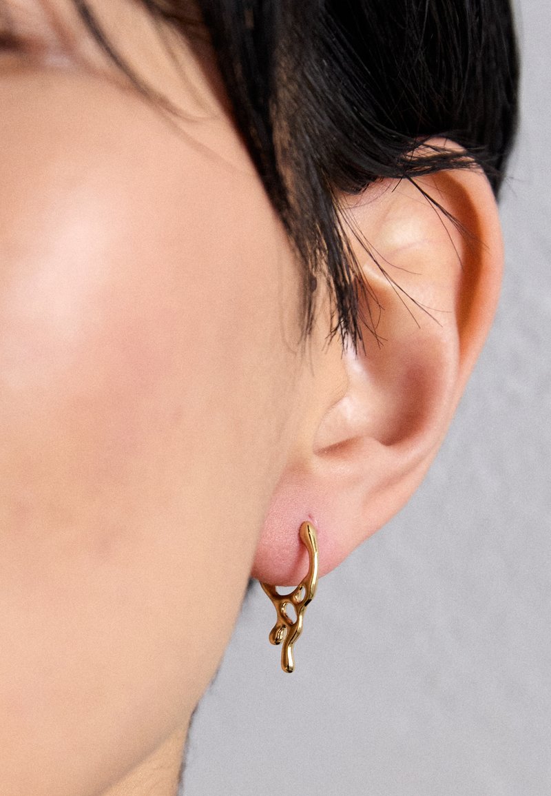 Boucle d'oreille en or avec un design organique et fluide, présentant des accents texturés et une finition lisse, portée sur une oreille percée.