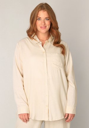 Vrouw met golvend rood haar, gekleed in een oversized crèmekleurig overhemd met lange mouwen en een voorzak, staand tegen een effen achtergrond.