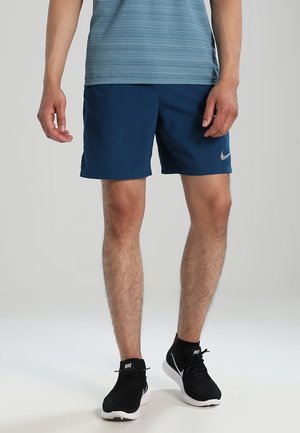 Mann trägt blaue sportliche Shorts, schwarze Nike-Sneaker und ein blaues, texturiertes Kurzarmhemd und steht vor einem schlichten Hintergrund.