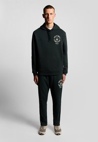 Donkergroene hoodie en bijpassende joggers van zachte stof, met een voortas en logodetail op de borst en dij.