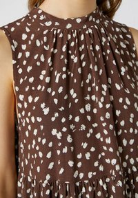 Brun ärmlös klänning med hög krage, med vita stänkta mönster och insamlade plisseringar. Tyget har en strukturerad yta.