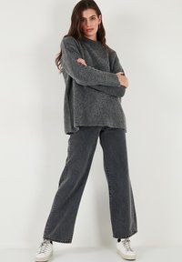 Pull en tricot gris avec une finition texturée, doté d'un col montant et d'une coupe ample, associé à un jean noir taille haute et des baskets blanches.