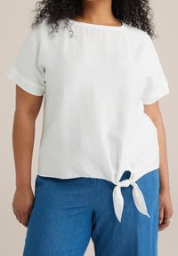 White, short-sleeve top with a wide neckline, featuring a knotted detail at the hem. Paired with blue, loose-fitting pants.