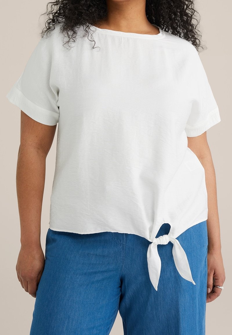 White, short-sleeve top with a wide neckline, featuring a knotted detail at the hem. Paired with blue, loose-fitting pants.