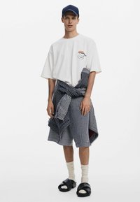 Tricou alb supradimensionat cu un grafic, pantaloni scurți în dungi bleumarin, cu același model așezat în jurul taliei, sandale negre și șosete albe.