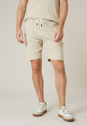 Man draagt beige cargoshorts met witte trekkoord en witte sneakers, staand tegen een effen lichte achtergrond.