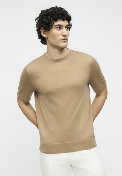 Νέος άνδρας με σγουρά σκούρα μαλλιά, φοράει μπεζ κοντομάνικο πουλόβερ και λευκό παντελόνι, στέκεται με τα χέρια πίσω από την πλάτη, κοιτάζοντας προς τα δεξιά.