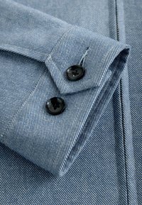 Gros plan sur le poignet d'une chemise en denim bleu clair pliée avec deux boutons noirs et une texture de tissu diagonale visible.