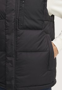 Schwarze Steppweste mit einem Reißverschlusstasche auf der Brust, gestepptem Muster und Seitentaschen. Verfügt über eine solide Konstruktion und einen abgerundeten Saum.