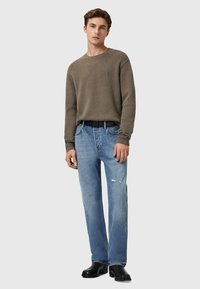 Olijfgroen trui met geribde manchetten, gecombineerd met lichtblauwe versleten denim jeans. Het model draagt een zwarte riem en zwarte schoenen.