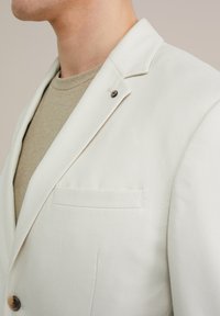 Weißer Blazer aus strukturiertem Stoff, mit einem Ein-Knopf-Verschluss und einer Brusttasche. Getragen über einem beigen Strickshirt.