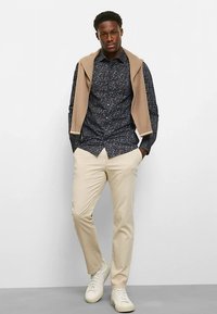 Marineblau gemustertes Hemd, beigefarbener Pullover lässig über die Schultern gelegt, cremefarbene Hose und weiße Sneaker. Texturierte Materialien, schlanker Schnitt.