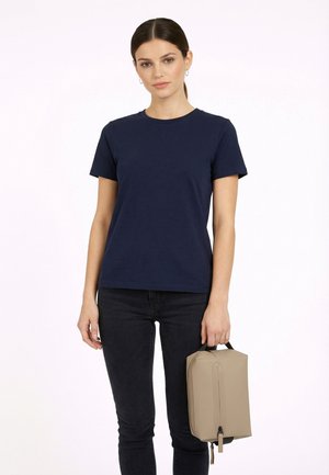 Mujer con camiseta azul marino y vaqueros negros sosteniendo una bolsa rectangular beige con asa contra un fondo blanco liso.