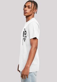 Vit bomullströja med svarta tryckta logotyper och grafik, korta ärmar och rundad halsringning. Gjord för avslappnad användning.