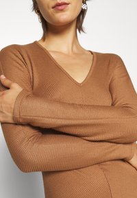 Maglia lunga marrone a maniche lunghe con scollatura a V, realizzata in tessuto texturizzato e con maniche aderenti, che mostra un aspetto liscio e elastico.