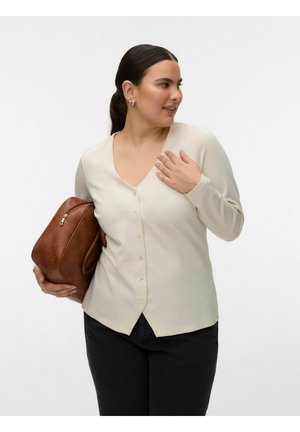 Sötét hajú nő krémszínű, gombos felsőben, aki barna bőr táskát tart az egyik karja alatt, oldalra néz, és a másik kezével megérinti a mellkasát.