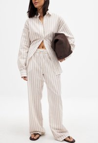 Beżowa i kremowa koszula z paskami na guziki oraz luźne dopasowane spodnie. Teksturowana tkanina z luźnym krojem. Trzyma brązową torbę w ręku.