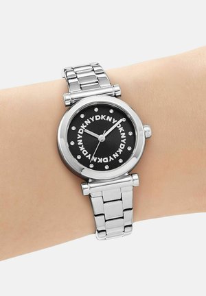 Montre-bracelet en métal argenté avec cadran noir, marqueurs d'heure en cristal, et logo "DKNY" répétitif au poignet affichant l'heure 2:09:36.
