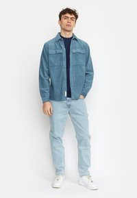 Giacca camicia in corduroy azzurro chiaro con due tasche sul petto, indossata sopra una maglia blu navy, abbinata a jeans azzurri chiari e sneakers bianche.