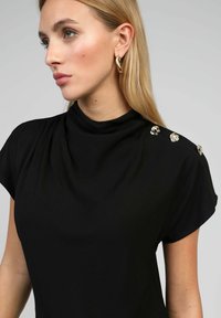Blouse noire avec un col montant et des manches courtes ; présente des boutons argentés décoratifs sur l'épaule, fabriquée en tissu lisse.