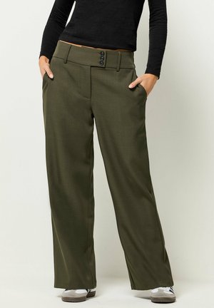 Osoba nosząca luźne oliwkowo-zielone spodnie z trzema czarnymi guzikami, czarny top z długim rękawem, białe trampki, ręce w kieszeniach, stojąca na białym tle.