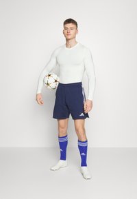 Langarmige witte sportshirt, marineblauwe shorts met witte accenten, blauwe sokken met witte strepen, een meerkleurige voetbals bal vasthoudend.
