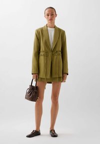 Green blazer with ruffled pockets, paired with a matching skirt. Brown drawstring bag and black flat shoes. White textured top underneath.