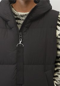 Svart pufferväst med hög krage, zippered front och fabric pull tab. Har en slät textur och subtil branding på remmen.