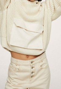 Kremowy sweter dzianinowy z dużą przednią kieszenią, zestawiony z wysokim stanem beżowych spodni z ozdobnymi guzikami i bocznymi kieszeniami.