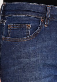 Mörkblå denimjeans med en synlig ficka, brun toppsöm och en mjuk textur. Har en klassisk femficksdesign.