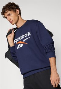 Námornícky modré mikina Reebok s bielym logom s oranžovým akcentom. Rybované manžety a lem, s hladkou textúrou. Model drží čiernu tašku.