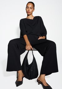 Top nero loose con scollo drappeggiato, pantaloni neri larghi e borsa nera con paillettes. Messo in evidenza da scarpe nere a punta con decorazioni a borchie.